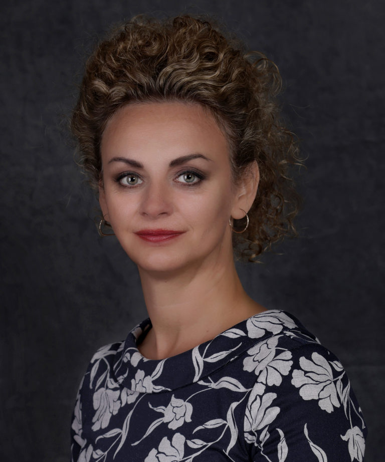 Lady with tied up curly hair wearing a blue & white flowery dress standing in front of a dark backdrop