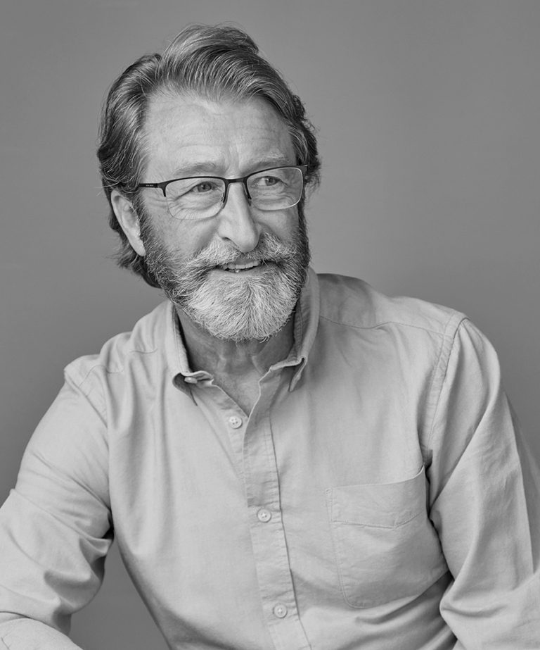 Man with beard smiling wearing an open neck shirt looking to one side whilst having his profile headshot taken in black & white.
