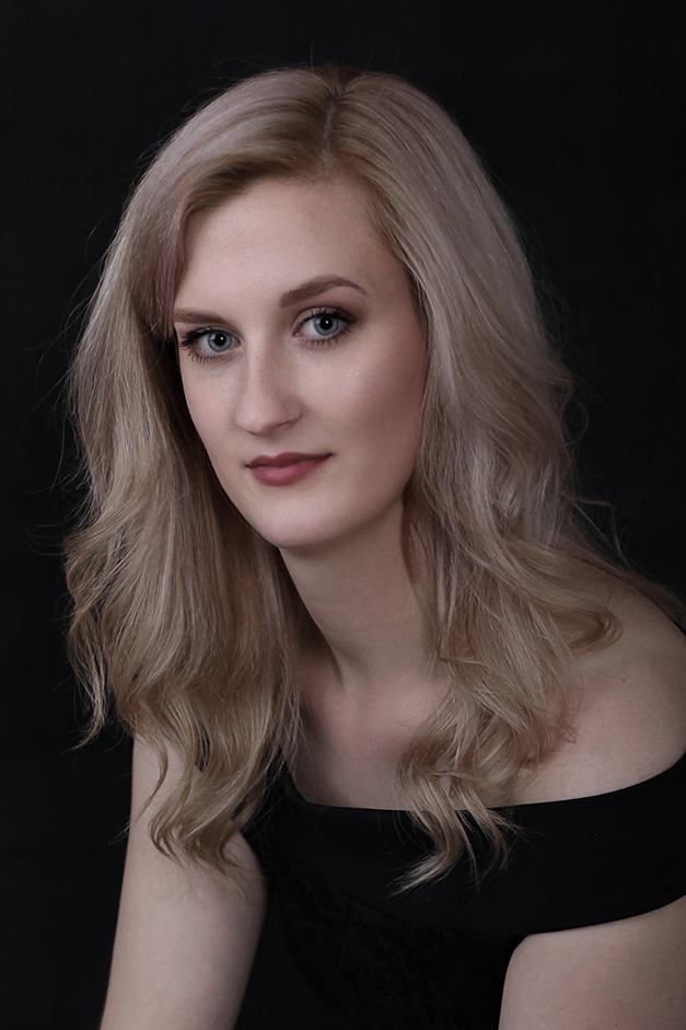 Young lady with blond hair and black off the shoulder dress sitting against a black backdrop