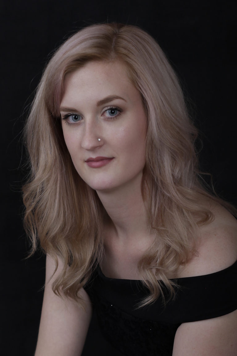 Young lady with blond hair and black off the shoulder dress sitting against a black backdrop