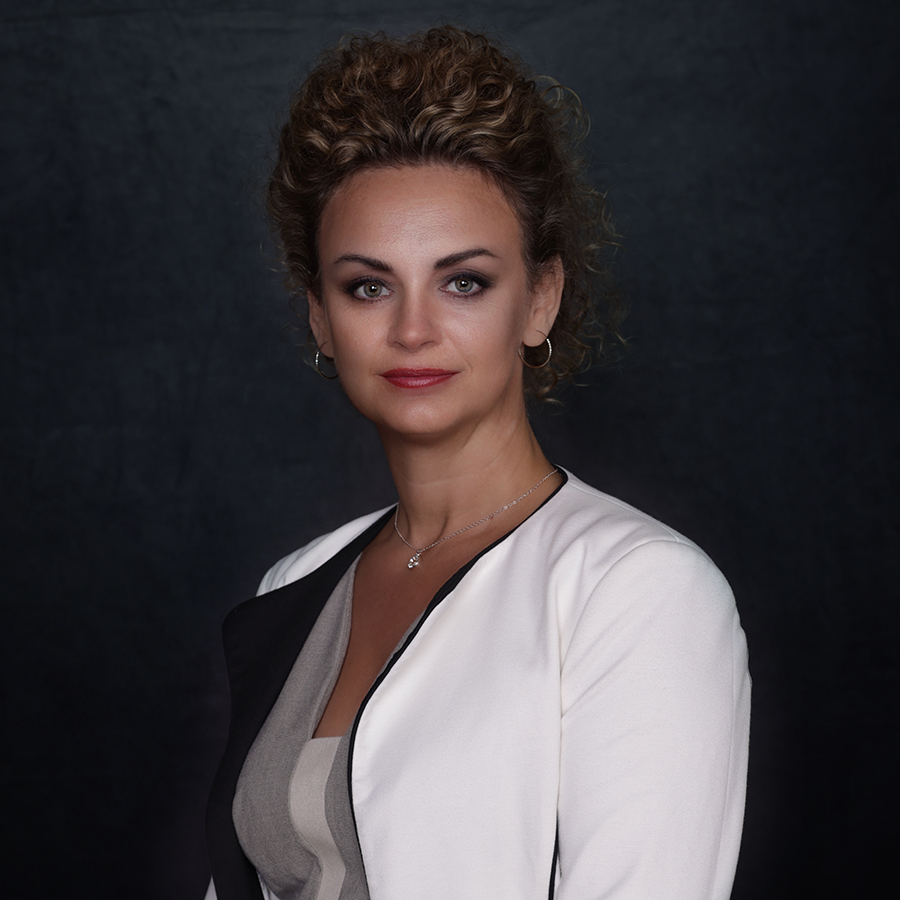 Lady with tied up curly hair wearing white business jacket and top standing in front of a dark backdrop