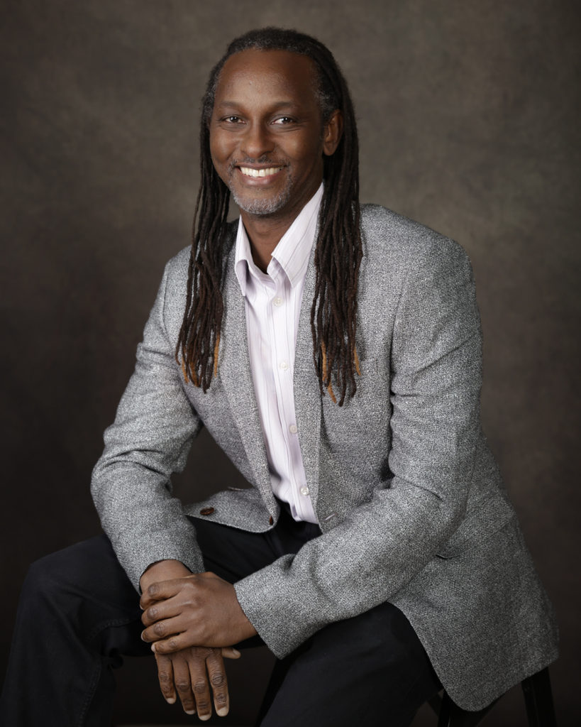 Black man wearing a suit jacket smiling for his LinkedIn headshot. The man has long dreadlock hair.