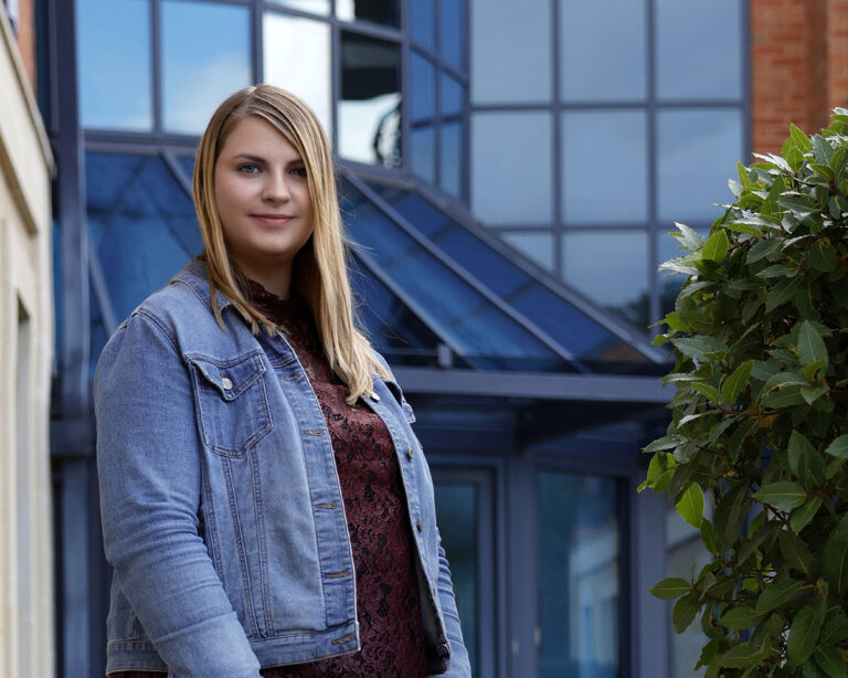 Business portrait taken outside a glass fronted office with blue tinted windows, the lady is wearing a blue denim jacket with a red blouse.